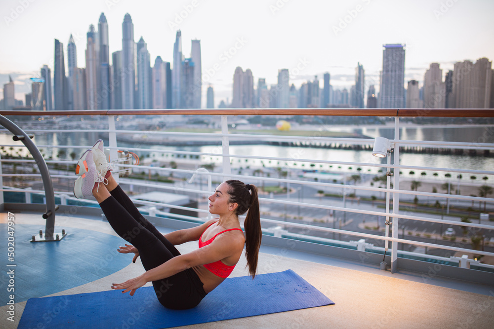 Young fit girl  on yoga mat in sport outfit doing abs workout on a on the outdoor in summer, , abs workout and hip joint mobility, enjoying morning aerobic classes with  panoramic view of city