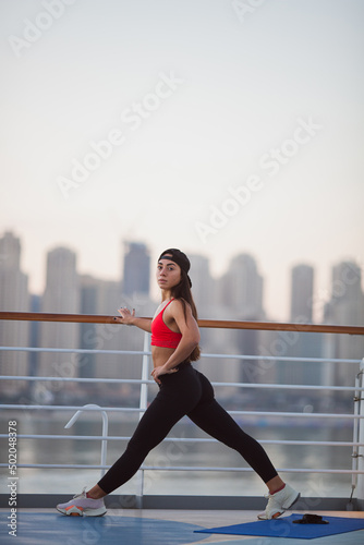 Fototapeta Naklejka Na Ścianę i Meble -  Sporty Healthy Lifestyle Concept. Sporty beautiful woman with long black hair doing legs stretching exercises, enjoying morning aerobic classes with beautiful panoramic view of city, morning work out
