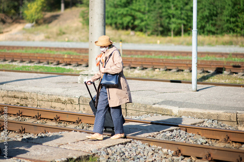 Elderly woman with suitcase and face mask crosses the railway tracks