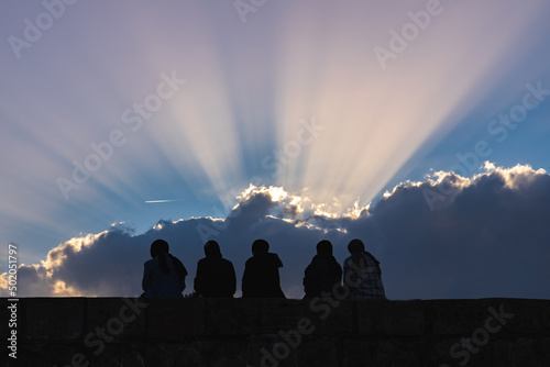 Silhouette of women wearing headscarves. Ray of light reflects from behind clouds at sunset. 