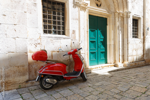 Red moped (scooter) is parked near the entrance to the building. Croatia, city of Trogir, sunny summer day. Paved road, beige walls. Selective focus, close-up.