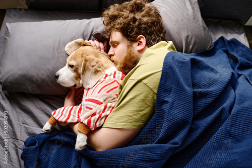 High angle view of bearded young man lying under blanket in bed and cuddling his dog photo