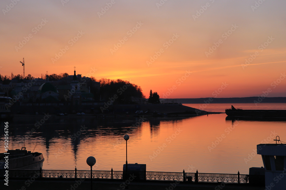 Silhouettes of the hilly old part of the city and the pier and reflections in the water against the background of a bright sunset.