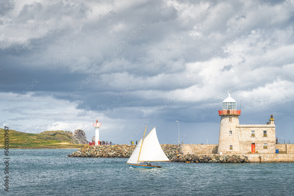 People on a small sailboat exiting Howth marina, passing Howth Lighthouse and per, with Irelands Eye island in the background, Dublin, Ireland