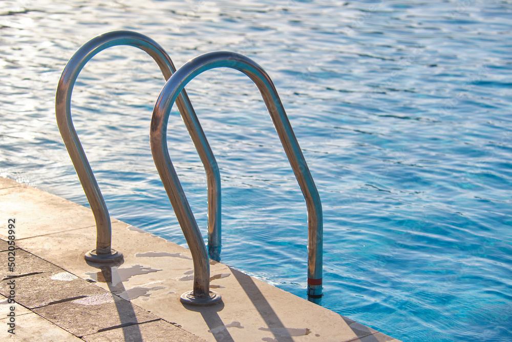 Close up of swimming pool stainless steel handrail descending into tortoise clear pool water. Accessibility of recreational activities concept