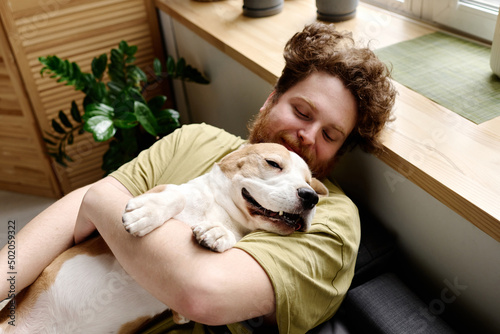 Young bearded man lying on bed and holding his sleeping dog on arms during rest time in bedroom photo