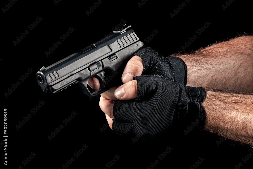 Closeup man hand with gloves holding gun with two hands and aiming down on black background. Finger on the trigger of the pistol. - obrazy, fototapety, plakaty 