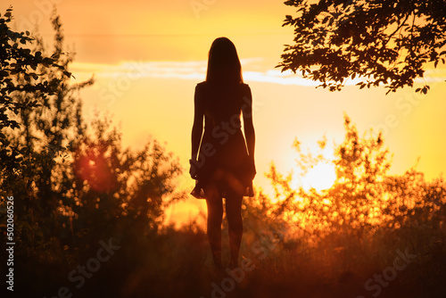Rear view of young woman in walking alone through dark woods at bright sunset. Nature exploration concept