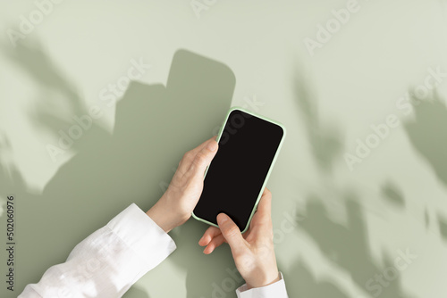 Mock up phone with black blank screen in female hands, white shirt sleeve, minimal flat lay on pastel green background. Summer lifestyle scene with smartphone, sunlight shadows, copy space photo