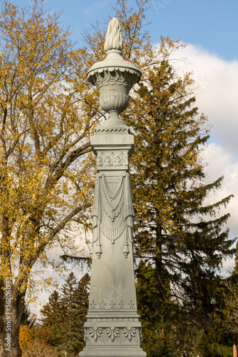 Tall zinc/white bronze gravestone with urn on top against yellow leaved trees photo