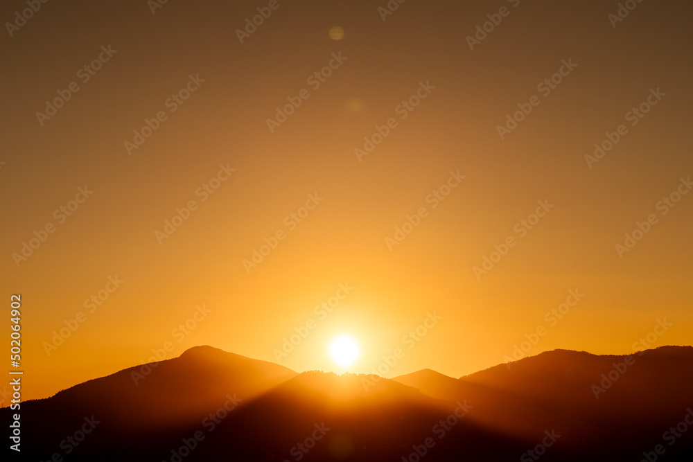rays of the sun setting behind a mountain with a completely clear, orange sky at sunset