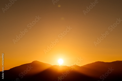 rays of the sun setting behind a mountain with a completely clear, orange sky at sunset