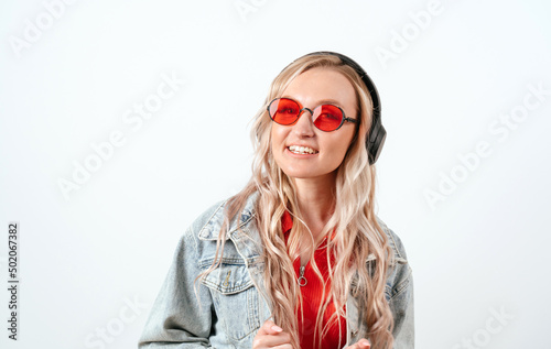 Blonde in headphones on a white background. She is wearing a denim jacket and red glasses. © cinematri