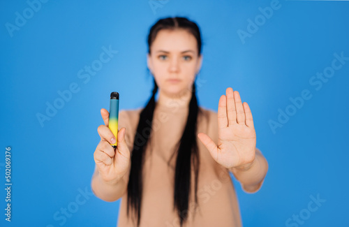 a girl on a blue background holds a disposable cigarette in one hand and shows a stop to smoking with the other hand. the concept of a healthy lifestyle