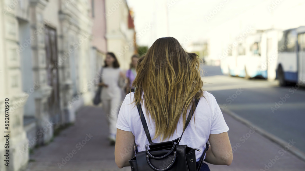 Rear view of a young girl in an urban street in the summertime. Media. Woman in white shirt with long hair walking on the background of the city.