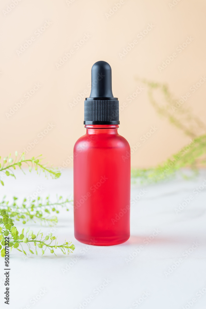 Red glass bottle with dropper pipette with serum or essential oil on marble table with green branches. Skin care cosmetic. Beauty concept for face body care Red glass bottle with dropper pipette with serum or essential oil on marble table with green branches. Skin care cosmetic. Beauty concept for face body care