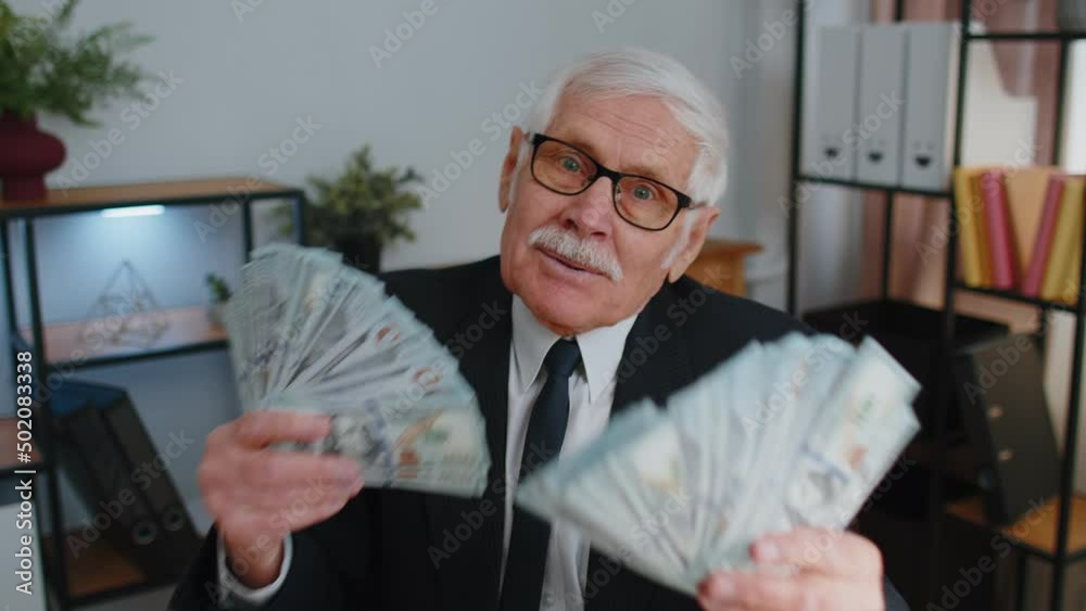 Senior mature banker business man accountant wears glasses, formal suit, celebrating business success dancing with stack of money dollar cash. Elderly grandfather freelancer indoors office interior