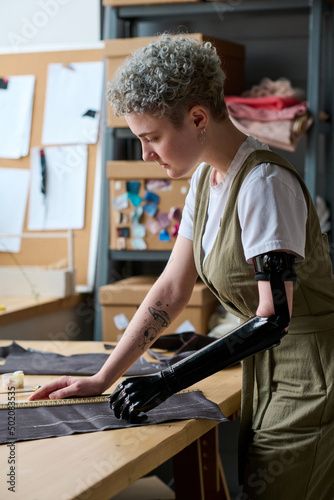 Young businesswoman with disability looking at piece of textile while standing by table and working over new fashion collection