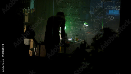 Rear view of stressed, tired young worker sitting at table, making fists in dim room. Stock. Exhausted young man working at night at a desk with many notes and photos, overwork concept.