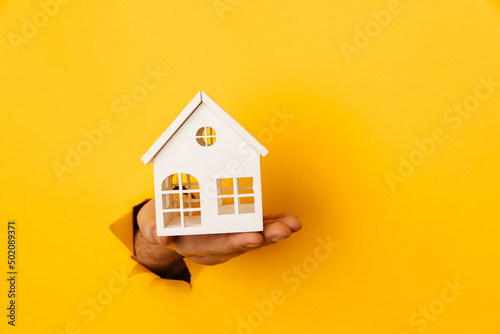 Male hand with mini house through a hole in yellow paper background
