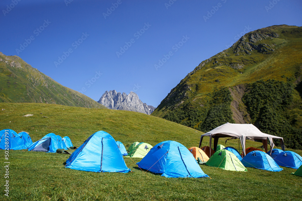 Tourist camping in the mountains of many tents and shelters for climbing to the top of the mountain - an alpine camp. Overnight in nature. Georgia, Chaukhi Mountain, Jute village
