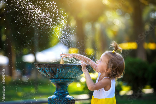 A funny little girl is playing with the spray of a drinking water fountain in the park. Childhood  child  summer time  heat. Solar protection for children s skin 