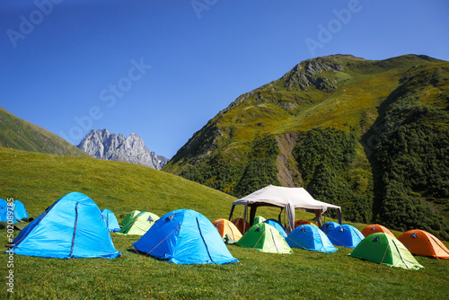Tourist camping in the mountains of many tents and shelters for climbing to the top of the mountain - an alpine camp. Overnight in nature. Georgia, Chaukhi Mountain, Jute village