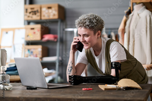 Happy young female tailor speaking on mobile phone with client while looking through new orders on laptop screen by workplace photo