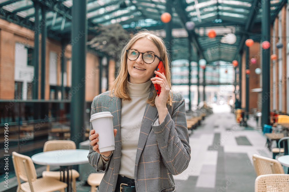 Fototapeta premium Happy beautiful businesswoman talking on cell phone outside, standing with coffee paper cup near coffee shop outdoors while walking in morning city. 
