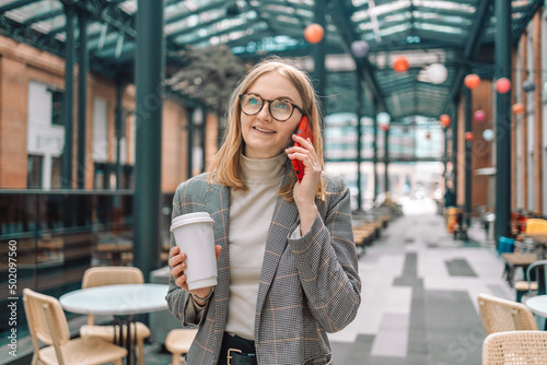 Happy beautiful businesswoman talking on cell phone outside  standing with coffee paper cup near coffee shop outdoors while walking in morning city. 