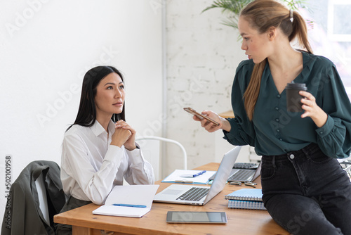 Two young women in the office. Talking, smiling, coffe break, pause. Diversity, asian female. Coffee break, chat and small talk with friends and colleagues, team members photo