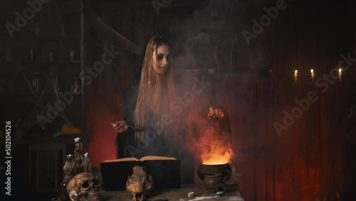 Halloween, witch use magic book and cauldron prepare glowing poison or love potion. Black magic occult female wizard in dark room cage spider web human skull Necromancer cooking magic potion. Handheld