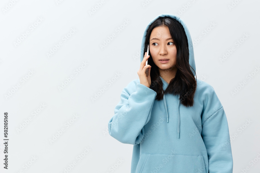Confused pretty Korean young female in blue hoodie hold phone get shock info in call posing isolated on over white studio background. The best offer for ad. Distance communication Gadget Ad concept Confused pretty Korean young female in blue hoodie hold phone get shock info in call posing isolated on over white studio background. The best offer for ad. Distance communication Gadget Ad concept