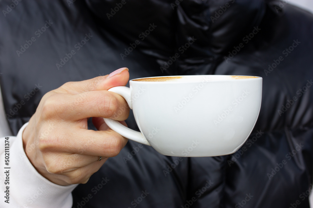 White porcelain coffee cup with hot cappuccino drink in woman's hand on dark background. Faceless girl drinking morning coffee outside in the afternoon. Human enjoying aroma caffeine beverage in cafe.