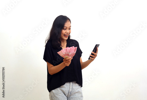 Happy asian young woman holding indonesian money and cellular phone. Isolated on white background