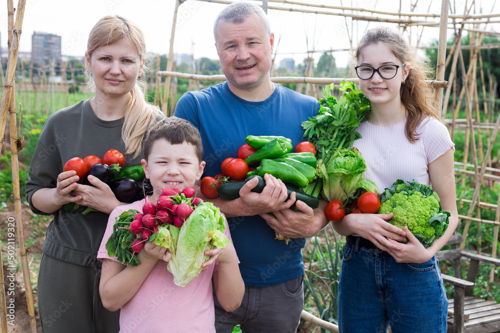 Portrait of happy family with two kids posing in garden with fresh harvest of vegetables and greens