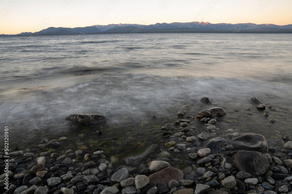 Long exposure shot of Nahuel Huapi lake at sunset. Beautiful blurred water effect, the rocky shore and Andes mountains in the horizon with a magical dusk light.