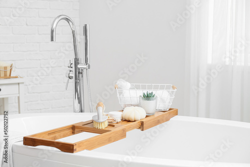 Wooden bathtub tray with body massage brush, cosmetic products and basket with towel in bathroom