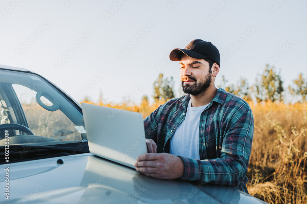 Young latin farmer man on his pickup truck and working on his laptop. Agricultural concept.