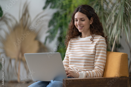 Joyful woman sitting on armchair relaxing while browsing online shopping website. Happy girl surfing net, chatting with friends during free time at cafe. Young female doing research work for business.