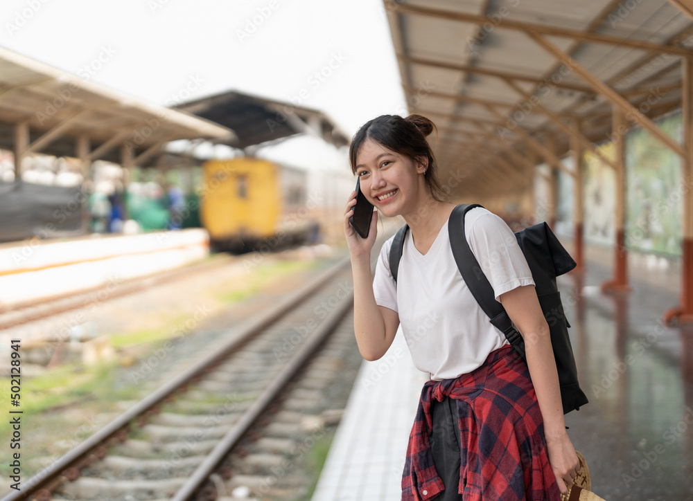 Beautiful young asian woman with a backpack uses the phone while standing near the railroad train on the platform. Cheap travel summer concept