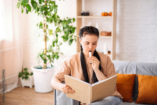 A beautiful young woman uses a disposable electronic cigarette on the couch at home reading a book. vaping concept