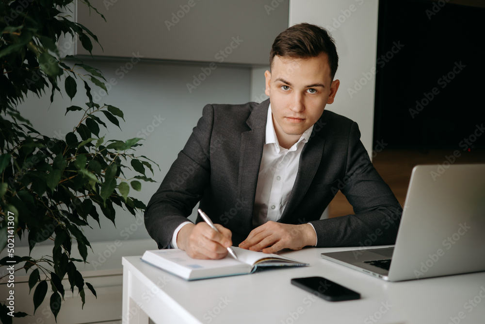 Serious young Caucasian man sitting at desk making notes in notebook Serious young Caucasian man sitting at desk making notes in notebook