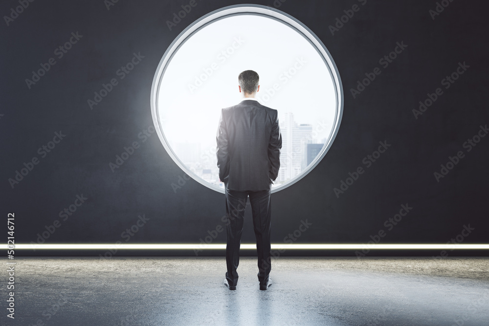 Obraz premium Back view of thoughtful young business man standing in creative modern concrete interior with round window and city view. Mock up place on wall and daylight.