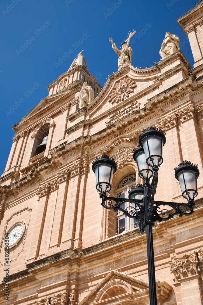 Facade of a big fabulous church in Malta with a street latern in front and a clear blue sky in the background. The letters on the front spell 'Jesus' in different languages. Facade of a big fabulous church in Malta with a street latern in front and a clear blue sky in the background. The letters on the front spell 'Jesus' in different languages.