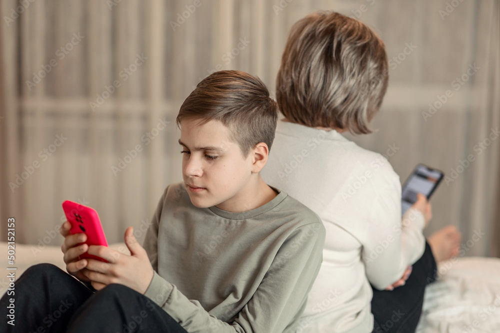 teenage son and mother sitting on the bed back to back and looking at smartphones, the concept of virtual communication in social networks, a waste of time and separation from the real world
