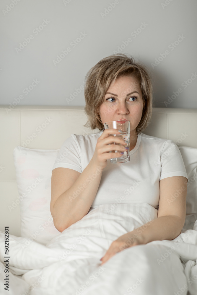 a middle-aged white woman with moisturized beautiful skin holds a glass of clean water in her hands, sitting on the bed. the concept of health and beauty care a middle-aged white woman with moisturized beautiful skin holds a glass of clean water in her hands, sitting on the bed. the concept of health and beauty care