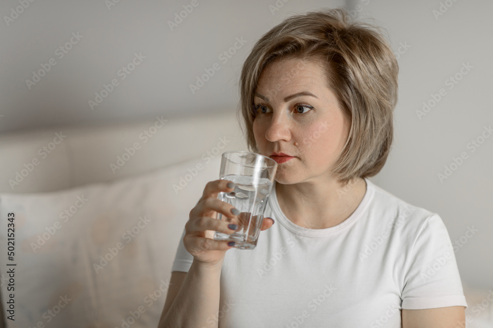 a middle-aged white woman with moisturized beautiful skin holds a glass of clean water in her hands, sitting on the bed. the concept of health and beauty care a middle-aged white woman with moisturized beautiful skin holds a glass of clean water in her hands, sitting on the bed. the concept of health and beauty care