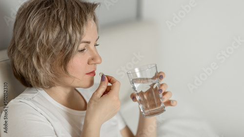 a middle-aged white woman with moisturized beautiful skin holds a glass of clean water and vitamins in her hands while sitting on the bed. the concept of health and beauty care