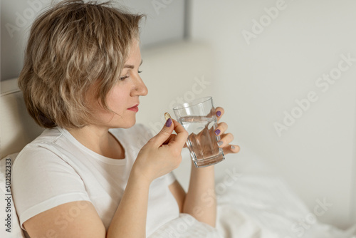 a middle-aged white woman with moisturized beautiful skin holds a glass of clean water and vitamins in her hands while sitting on the bed. the concept of health and beauty care